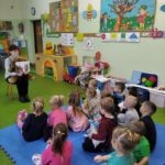 Przedszkolaki siedzą na dywanie i słuchają bajki czytanej przez bibliotekarkę