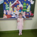Dziewczynka stoi na tle przymocowanej do tablicy planszy z szopką bożonarodzeniową Cicha noc