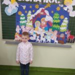 Chłopczyk stoi na tle przymocowanej do tablicy planszy z szopką bożonarodzeniową Cicha noc