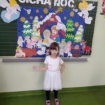 Dziewczynka stoi na tle przymocowanej do tablicy planszy z szopką bożonarodzeniową Cicha noc
