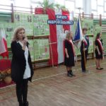 Narodowe Święto Niepodległości w SP 4 - wicedyrektor szkoły w udekorowanej sali przemawia przez mikrofon, dalej stoi poczet sztandarowy szkoły