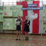 Narodowe Święto Niepodległości w SP 4 - dwie uczennice stoją na tle oklicznościowej dekoracji, jedna z nich czyta