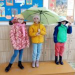 Jedna dziewczynka trzyma parasol, a inne dzieci ubrane są w ciepłe czapki, kurtki i gumowe kalosze